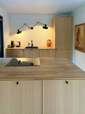 Une cuisine moderne avec des meubles en bois, un plan de travail, un évier, une bouilloire, une machine à café et un luminaire suspendu.