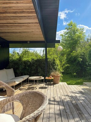 Une terrasse en bois avec un espace salon couvert, une table ronde et une plante en pot.