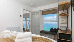 A bedroom with a bed, a bench, a window with a view of the sea, a desk, and a hanging garment rack. - The Observatory | Boutique Coastal Retreat | Panoramic Sea Views | Beach 100m (St Ives)