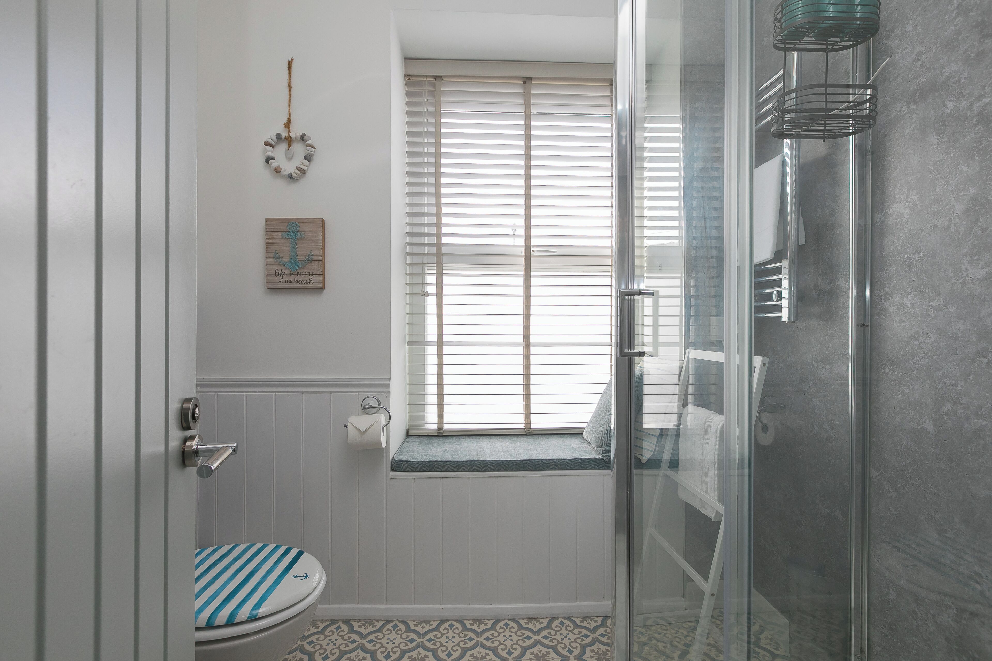 A bathroom with a toilet, a window with blinds, a shower area, and a small rug on the floor.