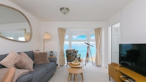 A living room with a sofa, a small table, a television, and a view of the sea through a bay window. - The Observatory | Boutique Coastal Retreat | Panoramic Sea Views | Beach 100m (St Ives)