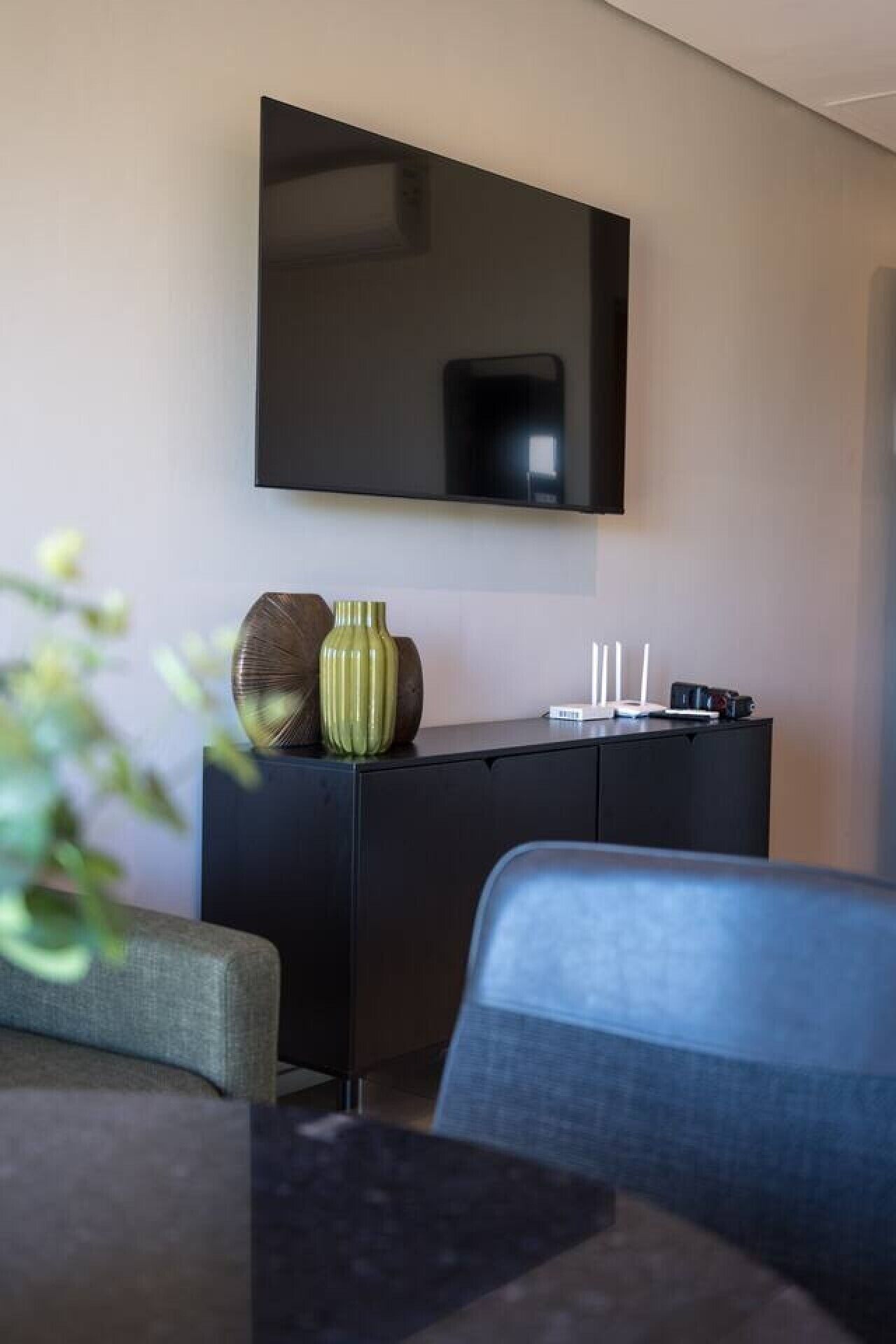 Une chambre moderne avec un téléviseur à écran plat fixé au mur, un meuble de couleur sombre agrémenté d’objets décoratifs et un canapé gris.