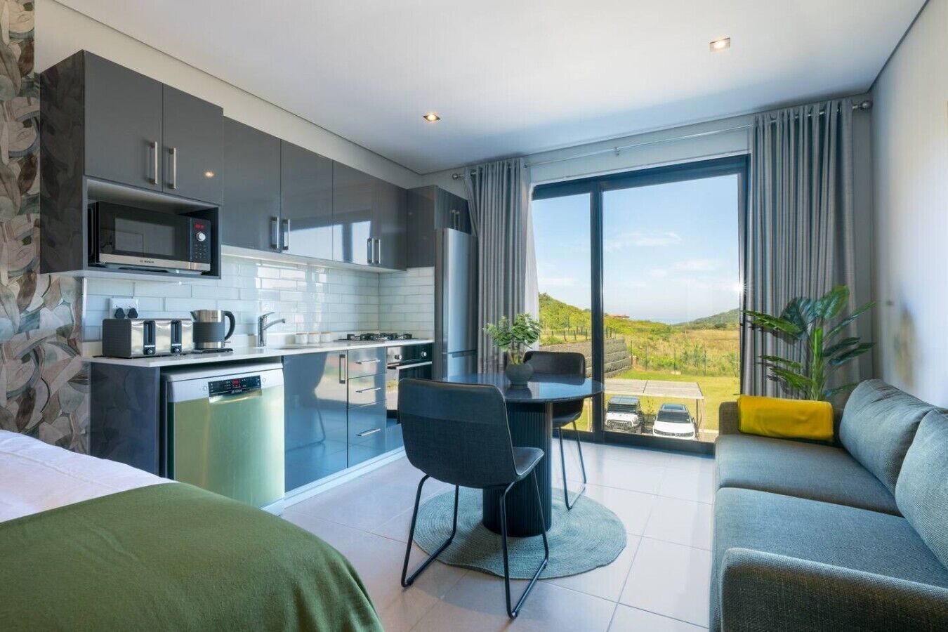 A modern hotel room with a kitchenette, a dining area, a sofa, and a large window offering a view of the countryside.
