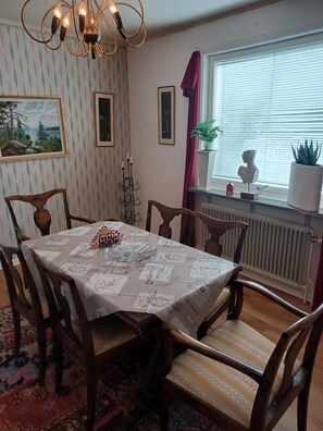 Un coin repas avec une table dressée pour quatre personnes, entourée de chaises en bois. Un radiateur se trouve sous la fenêtre, et un pot de fleurs est posé sur le rebord de la fenêtre.