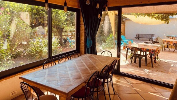A dining area with a large table and chairs, large windows with curtains, and an outdoor seating area visible through the windows. - The Cocon Lou Jaïa (Corbère)