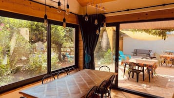 Een eetruimte met een grote tafel en stoelen, grote ramen voorzien van gordijnen, en een zichtbare buiten zitplaats.