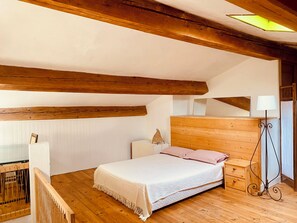 Une chambre avec un plafond en bois, un lit avec un matelas blanc et des oreillers roses, une table de chevet avec une lampe, et une petite armoire en bois.