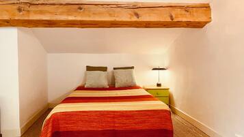 Een slaapkamer met een bed, twee kussens, een nachtkastje met een lamp en zichtbare houten balken in het plafond.