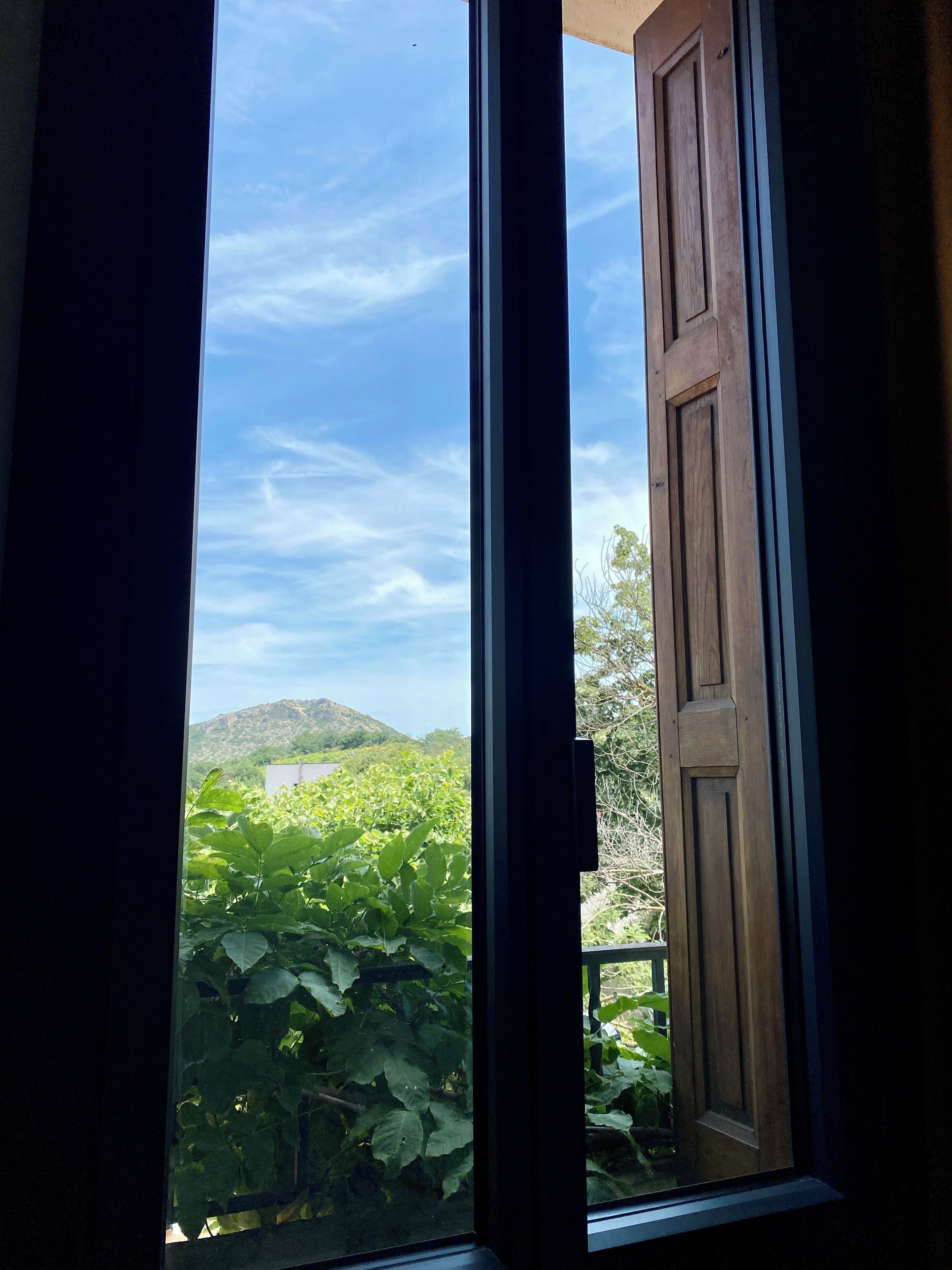Une vue à travers une porte ouverte, dotée d’un cadre en bois, qui donne sur un balcon où la végétation luxuriante contraste avec une colline lointaine.