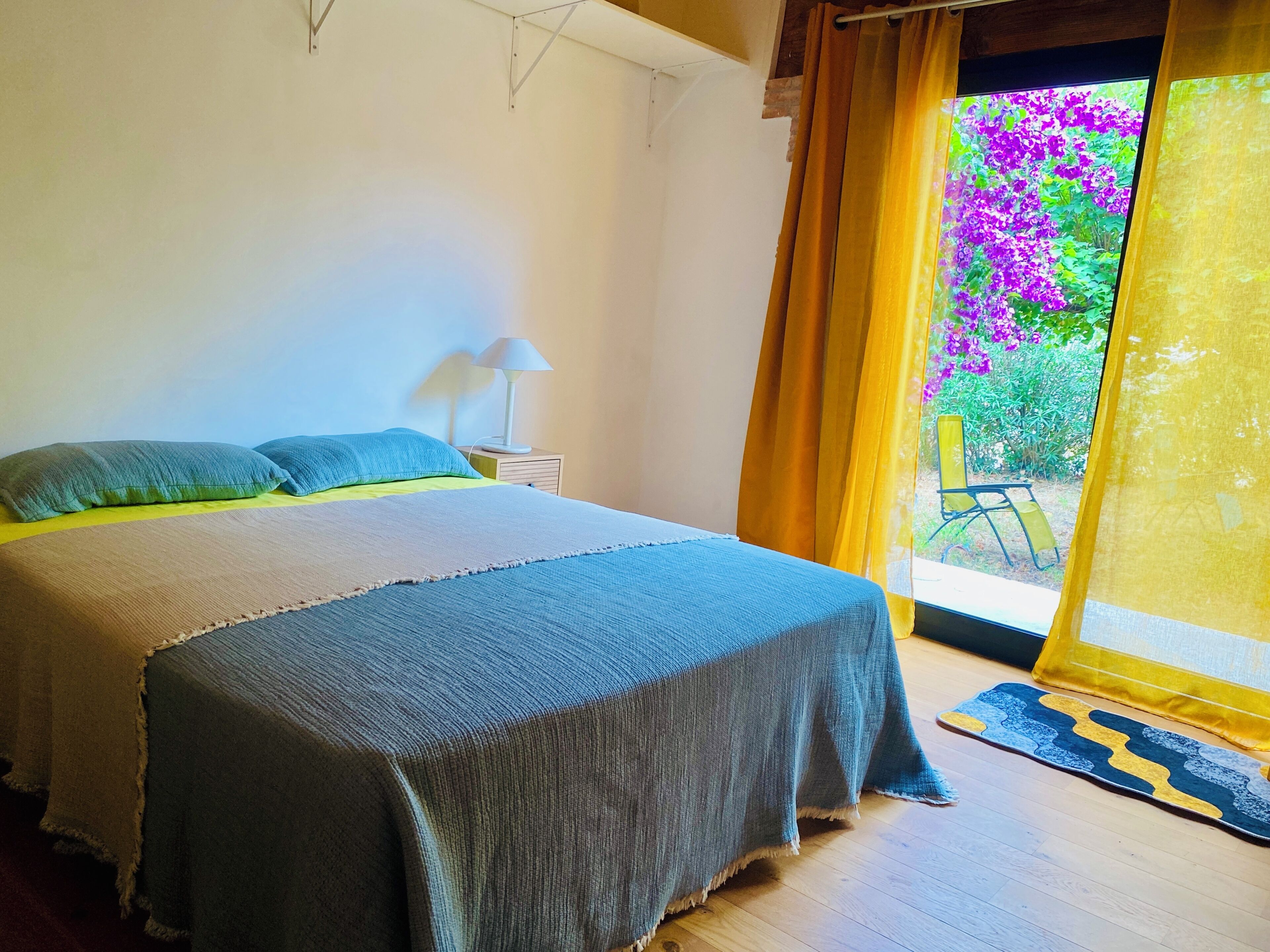 Une chambre à coucher avec un lit, une table de chevet, une lampe, une fenêtre avec des rideaux et une vue sur la verdure à l’extérieur.