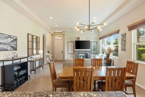 A spacious dining area with a wooden table and chairs, a modern chandelier, and a kitchen in the background. - Sky Villa Puerta Azul Retreat w Pools Gym (La Quinta)