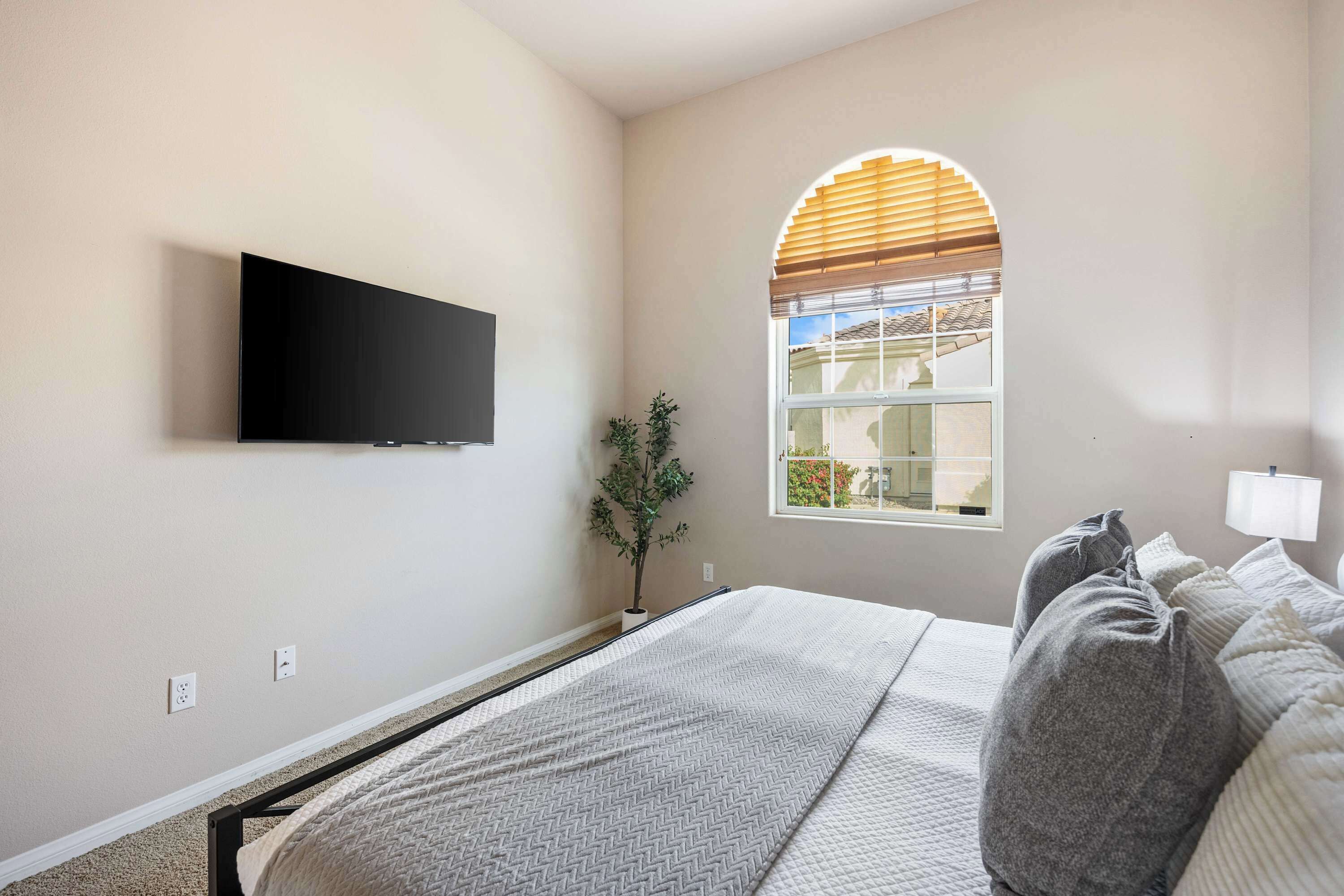 A bedroom with a bed, a TV mounted on the wall, a window with blinds, and a potted plant.