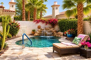 A pool with a waterfall, surrounded by palm trees and cacti, with a lounge chair and a sun dial. - Casa Veranda Puerta Azul Pool Retreat (La Quinta)