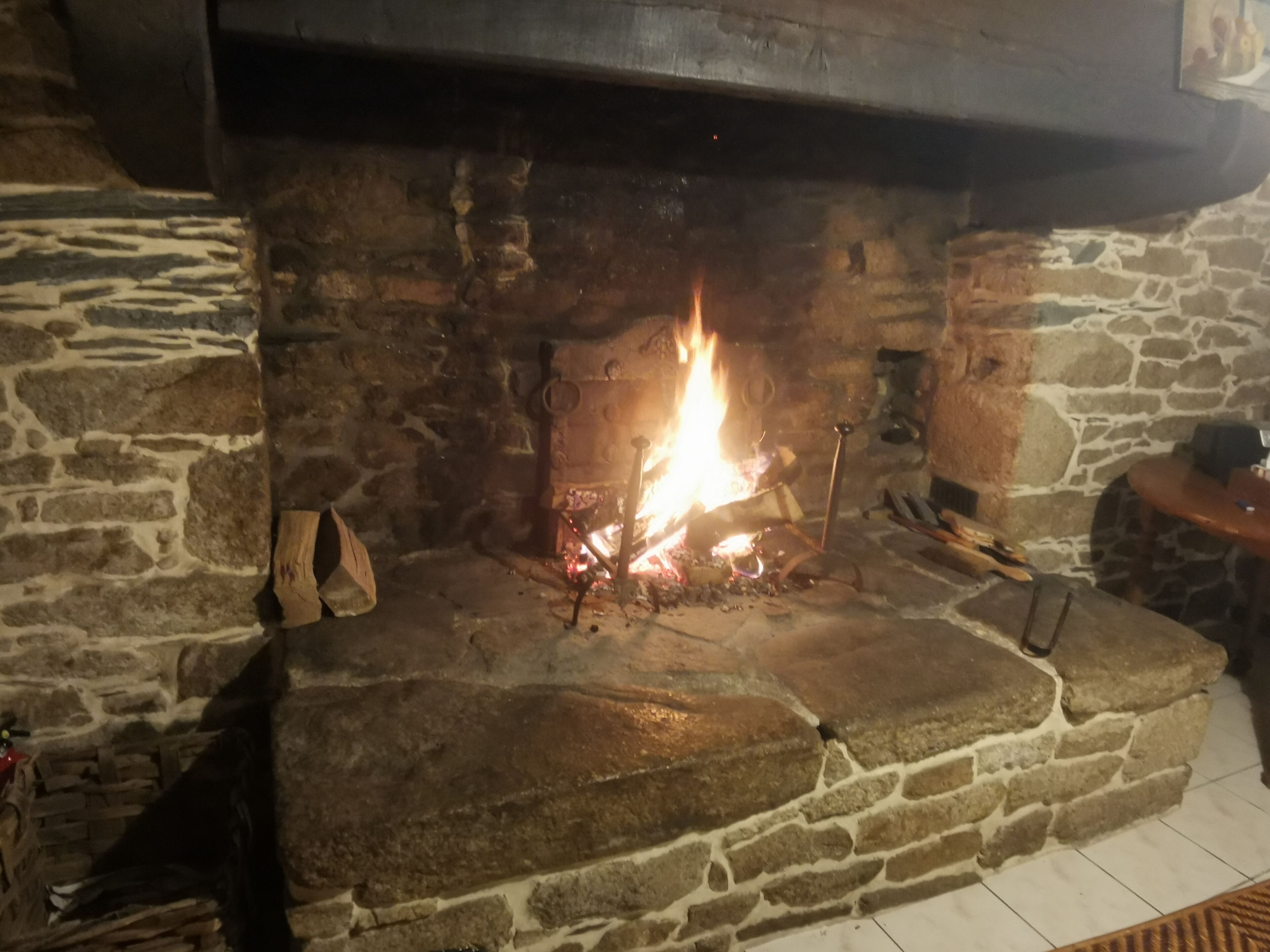 Una chimenea de piedra con fuego encendido, un banco de madera a la derecha y una pared de piedra.