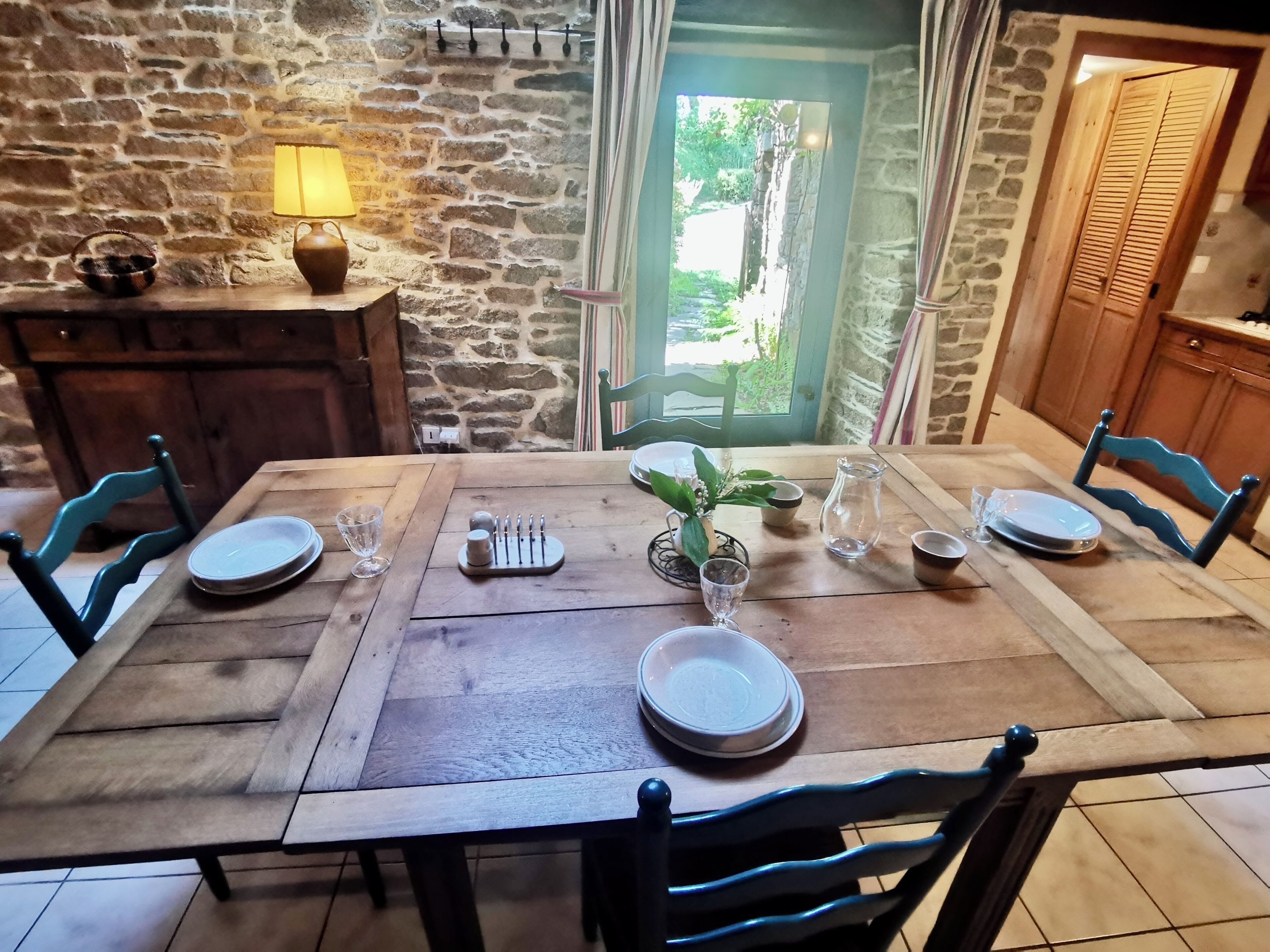 Una mesa de comedor dispuesta con platos, vasos y una jarrón, rodeada de sillas, en una habitación con paredes de piedra y una puerta de madera.