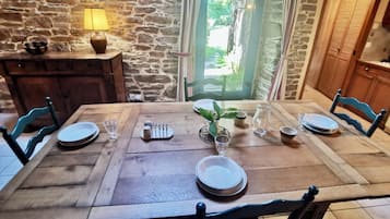 Una mesa de comedor dispuesta con platos, vasos y una jarrón, rodeada de sillas, en una habitación con paredes de piedra y una puerta de madera.