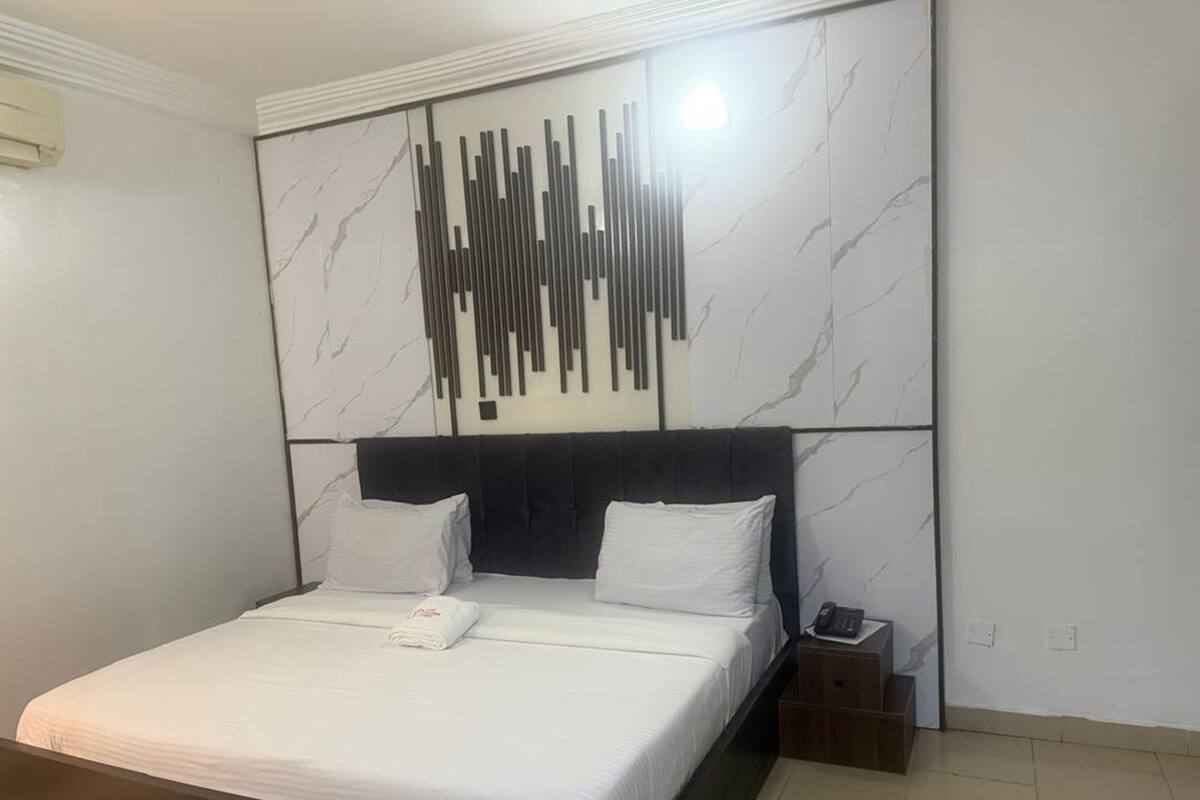 A modern bedroom with a large bed, a headboard, a wall-mounted light, and a bedside table with a phone.