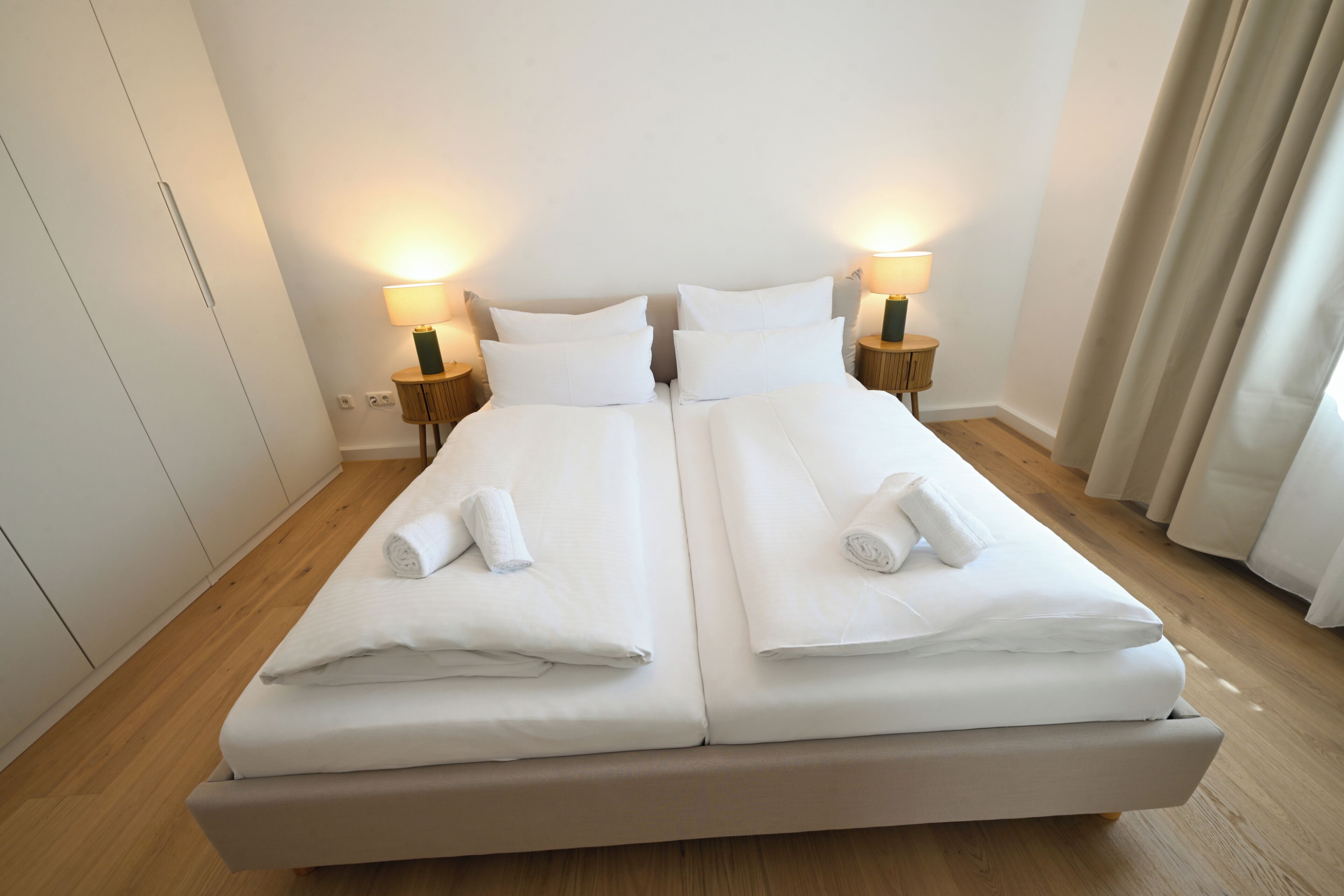 Ein Doppelbett mit weißen Bettwäsche und Handtüchern, zwei Nachttische mit Lampen und Holzboden.