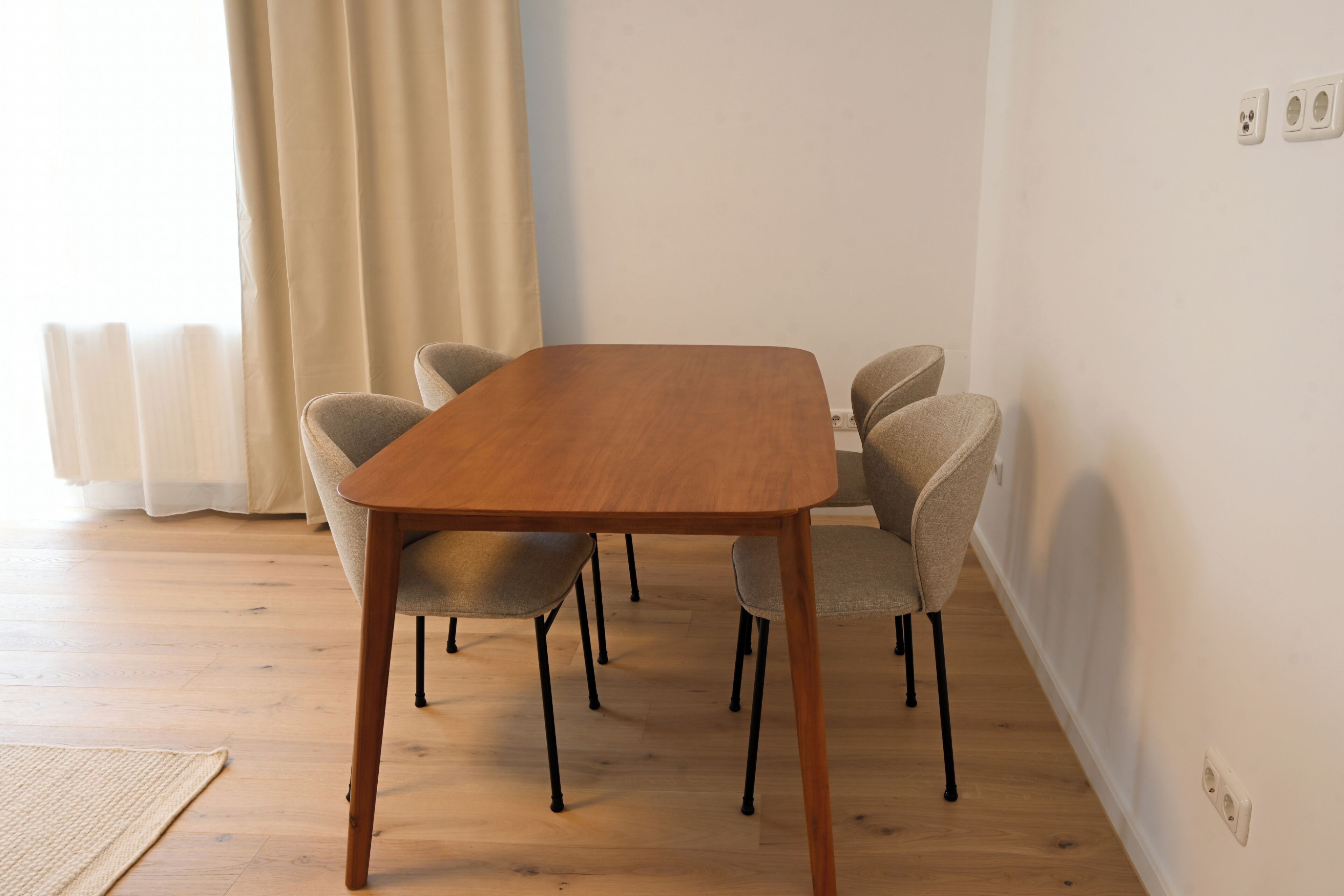A dining area with a wooden table and four chairs, a window with curtains, and a radiator.