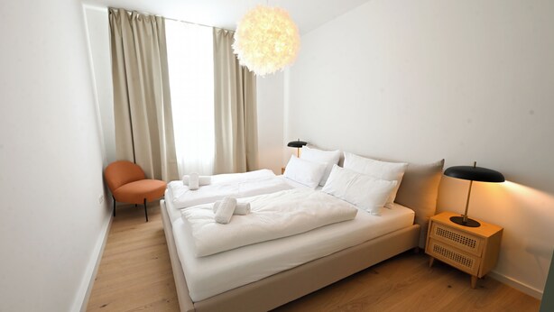 A double bed with white linens, a side table with a lamp, a chair, and a large window with curtains. - Liboria vacation apartment Starnberg with balcony for 6 persons (Starnberg)