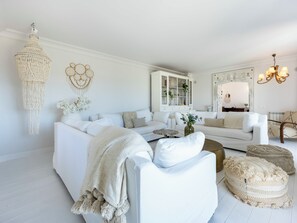 A white living room with a sofa, armchairs, a coffee table, a macrame wall hanging, a cabinet, and a chandelier.