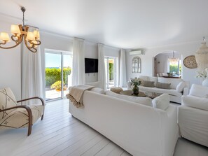 A spacious living room with a white sofa, a wooden chair, a round coffee table, and a chandelier. - Casa Paraíso - Sea View Villa in Praia da Luz (Luz)