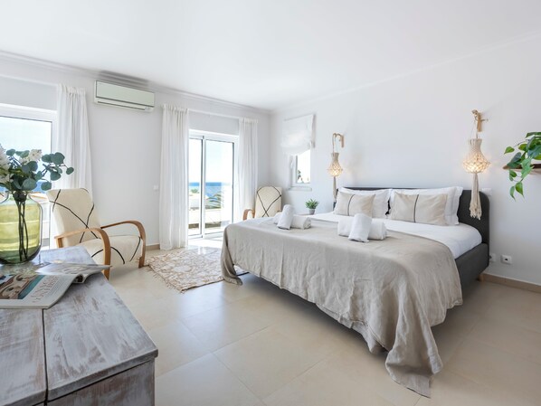 A bedroom with a bed, a chair, a table with a magazine, and a view of the sea through a sliding door.