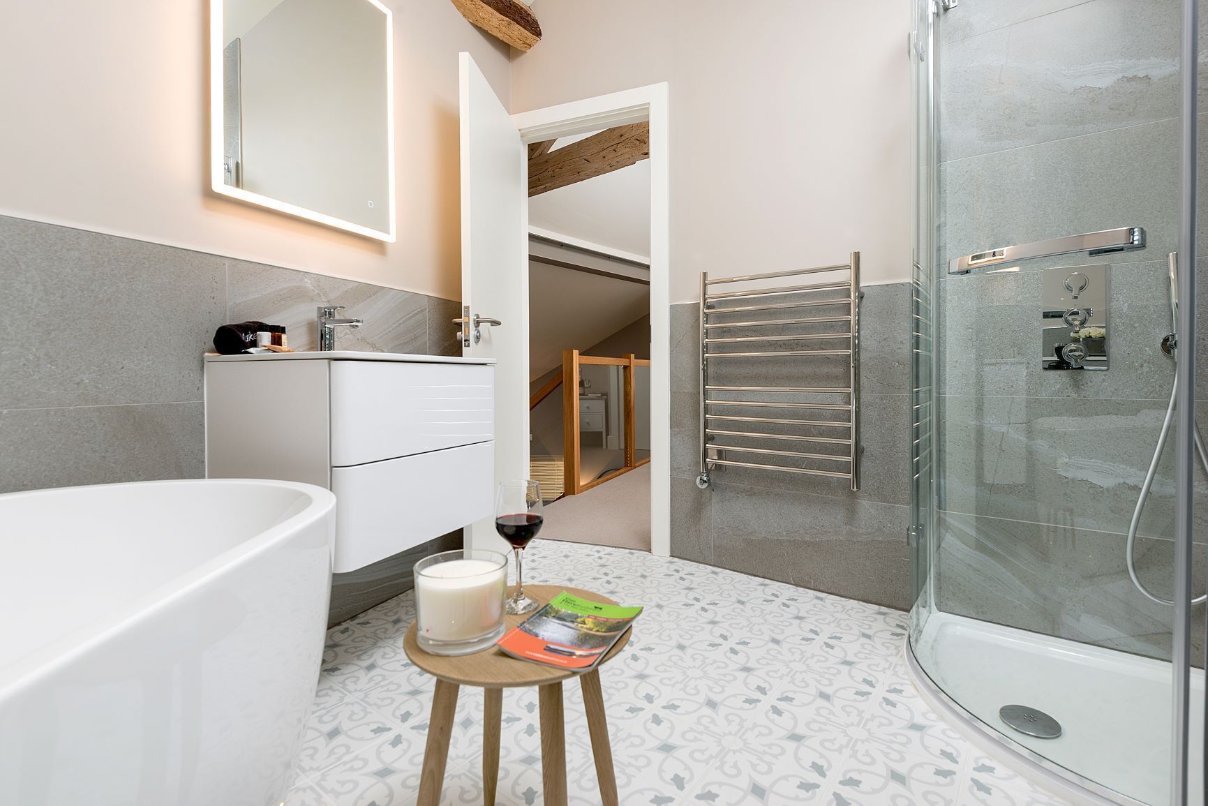 A modern bathroom with a bathtub, a glass shower enclosure, a towel rack, and a small table with a candle and a glass of wine.