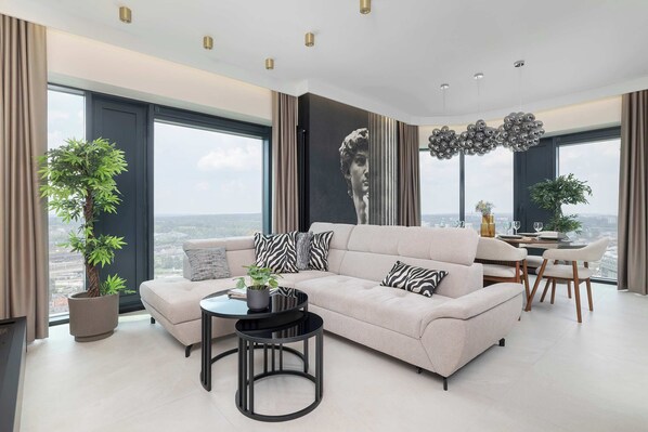 A modern living room with a sofa, coffee table, and dining area, large windows, and a city view. - Luxury Apartment with Air Conditioning | 25th Floor (Katowice)