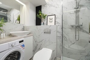 A modern bathroom with a marble wall, a sink, a toilet, a washing machine, and a shower. - Bright Apartment in the City Centre | Family City Break (Łódź)