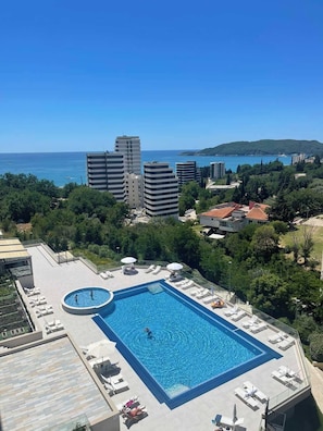 Ein Dachpool mit Liegestühlen und Sonnenschirmen, umgeben von Grünflächen und Gebäuden, mit Blick auf ein Gewässer.