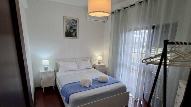 Habitación de hotel con cama, lámparas de noche, una lámpara colgante, una ventana con cortinas y un perchero.