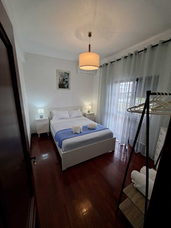 A hotel room with a bed, bedside lamps, a hanging light fixture, a window with curtains, and a coat rack. - Constança House 10 min. from Fonte da Telha (Corroios)
