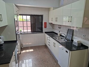 A modern kitchen with white cabinets, a black countertop, a built-in dishwasher, and a window with a view of the outside. - Constança House 10 min. from Fonte da Telha (Corroios)
