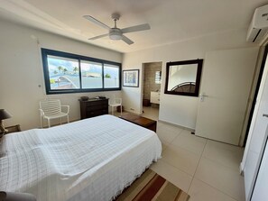 A bedroom with a bed, a ceiling fan, a window with a view, a bathroom with a mirror, and a door. - The Marina Suites (Saint francois)