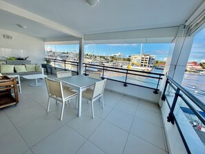 En balkon med udsigt over både og bygninger, et spiseområde med stole og bord, samt en sofa med puder.