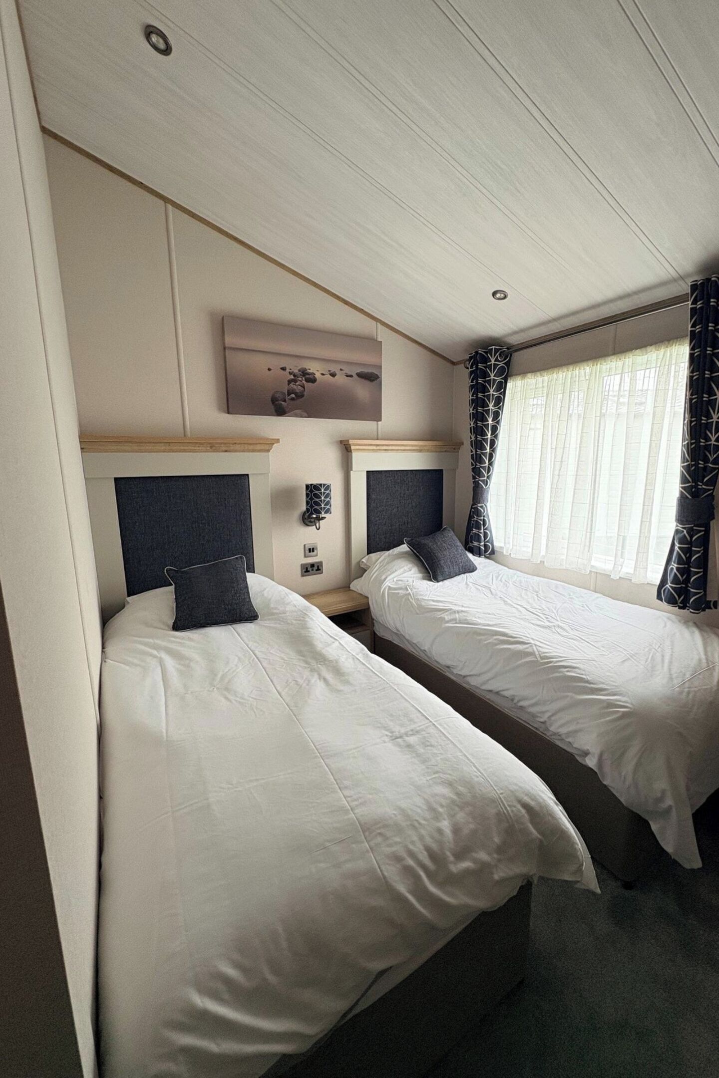 Ein Hotelzimmer mit zwei Betten, einem Kopfteil, einem Nachttisch, einer Wandlampe und einem Fenster mit Vorhängen.