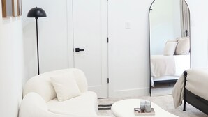 Une chambre à coucher moderne et minimaliste, avec un grand canapé blanc, une petite table basse blanche, un lit doté d’une tête de lit blanche, un grand miroir et un lampadaire noir.