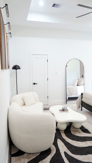 Ein modernes, minimalistisches Schlafzimmer mit einer großen weißen Couch, einem kleinen weißen Couchtisch, einem Bett mit weißem Kopfteil, einem großen Spiegel und einer schwarzen Stehlampe.