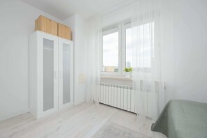Ein Zimmer mit Heizkörper, einem Fenster mit durchsichtigen Vorhängen und einem Bett mit einer grünen Tagesdecke.