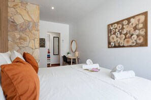 Hotellihuone, jossa on yksi seinä koristeltu kivellä, valkoisilla tekstiileillä varustettu sänky, työpöytä tuolilla, peili ja sieniä esittävä taulu.