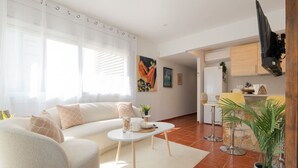 Sala de estar com sofá, mesa de centro e televisão. Há uma área de cozinha com balcão e mesa de jantar.