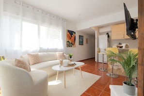 Ein Wohnzimmer mit Sofa, Couchtisch und Fernseher. Es gibt eine Küchenzeile mit Bar und Esstisch.