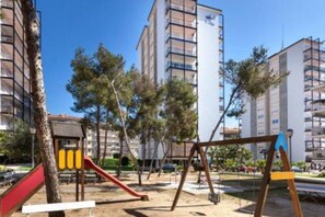 Ein Spielplatz mit Schaukeln und Rutschen, umgeben von Bäumen und hohen Gebäuden.