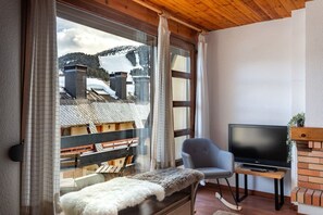 Une pièce avec une télévision, un fauteuil, un plaid en fourrure et une vue sur les bâtiments par la fenêtre.