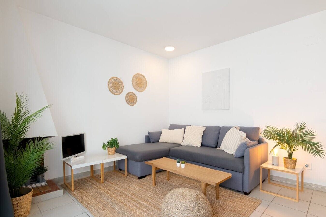 Una sala moderna con un sofá seccional gris, un televisor de pantalla plana, una mesa de centro de madera y dos mesitas de noche con plantas en macetas.