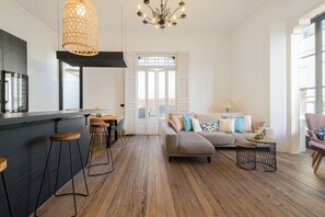 Ein modernes Wohnzimmer mit einer Couch, einem Essbereich mit Hockern, einer Küche mit Bar und einem großen Fenster.