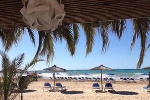 Uma praia com fileiras de espreguiçadeiras e guarda-sóis, palmeiras e um céu azul límpido.