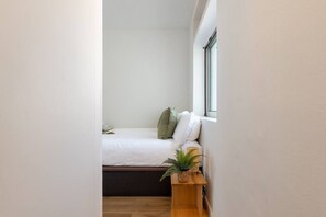 Een knus, netjes ingerichte slaapkamer met een bed, kussens, een nachtkastje met een plant en een raam.