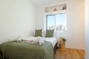 Un dormitorio con una cama, almohadas, un ventanal, una mesita de noche con una planta y vistas a edificios.